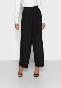 Široké černé kalhoty z lehkého materiálu, s předními plisovanými detaily a vysokým pasem. Nosené s fitted bílou košilí s dlouhým rukávem a černými sandály.