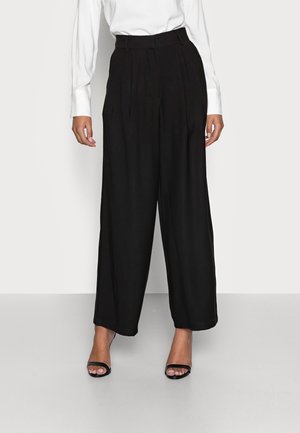 Weite schwarze Hosen aus leichtem Stoff, mit vorderen Falten und hoher Taille. Kombiniert mit einem eng anliegenden weißen Langarmhemd und schwarzen Sandalen.