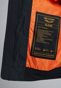 Sort jakke med orange for, der har et etiket med produkt-specifikationer, herunder materialer, lomme-design og pasformdetaljer.