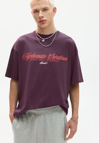 Lilla bomulds T-shirt med korte ærmer, med rød tekst "Hyphenate Créateur" på forsiden. Kombineret med grå shorts og sølv kæder.