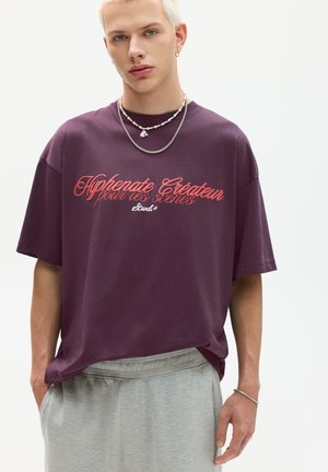 Lilla bomulds T-shirt med korte ærmer, med rød tekst "Hyphenate Créateur" på forsiden. Kombineret med grå shorts og sølv kæder.
