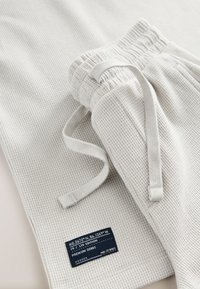 Baltos spalvos, dryžuotas trikotažinis sportinės kelnės su raišteliu juosmenyje, ant kurio siūtas lipdukas su koordinatėmis ir leidimo informacija.