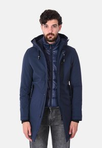 Parka blu navy con cappuccio e cerniera frontale, indossata sopra un gilet imbottito blu. Presenta tasche laterali e una superficie liscia.