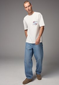 White short-sleeve t-shirt with a chest pocket featuring colorful graphic design, paired with loose-fit light blue denim jeans and brown slip-on shoes.