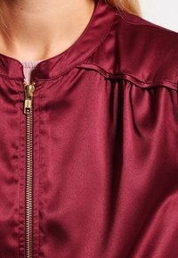 Burgundy satin bomber jacket with a gold zipper, round collar, and gathered detailing at the shoulders. Smooth, glossy texture.