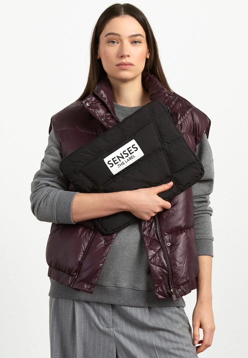 Puffer vest in deep purple with a glossy finish, worn over a gray sweater, holding a black quilted pouch labeled "SENSES THE LABEL."