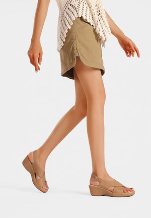 Donna che cammina indossando sandali beige con zeppa, mini gonna oliva con spacco laterale e top bianco senza maniche in crochet su sfondo semplice.