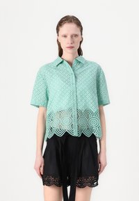 Mintgroene shirt met korte mouwen, met oogjespatronen en een gegolfde kantzoom. Draag het over zwarte shorts met kantafwerking.