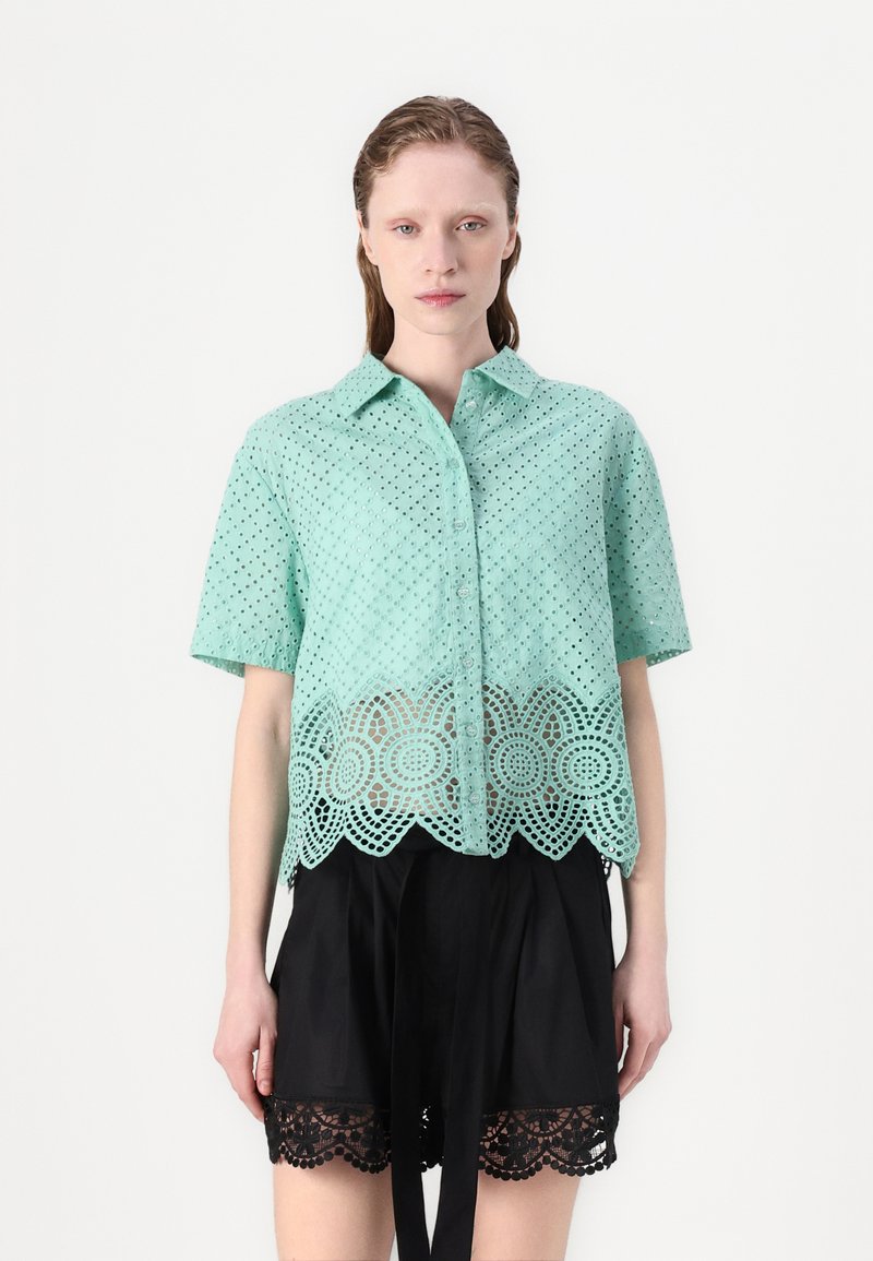 Mintgroene shirt met korte mouwen, met oogjespatronen en een gegolfde kantzoom. Draag het over zwarte shorts met kantafwerking.