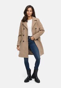 Cappotto beige doppiopetto con grandi bottoni neri, tasche laterali, rever a scollo, indossato sopra una maglietta bianca e jeans blu, abbinato a stivali neri.
