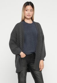Chunky strikkegenser i mørkegrått, åpen frontdesign, ribbestrikkede mansjetter og teksturert mønster. Brukt over en marineblå topp og svarte bukser.