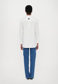 Weißes Langarmhemd mit Rückenschlitz und schwarzem Logo am Kragen, kombiniert mit blauen Denim-Hosen mit runder Patte.