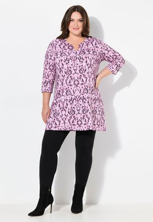 Rosa Tunika mit schwarzem und lila abstraktem Muster, 3/4-Ärmel, V-Ausschnitt, kombiniert mit schwarzen Leggings und Stiefeletten.