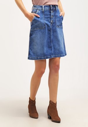Femme portant une jupe en denim bleu à boutons sur le devant avec de grandes poches avant et des bottines en daim marron, debout les mains dans les poches.