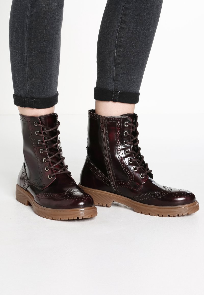 Botas de tobillo de cuero burdeos brillante con detalles de brogue, cordones en la parte delantera, cremallera lateral y una suela de goma marrón texturizada.