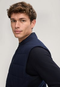 Granatowa pikowana kamizelka z wysokim kołnierzem i zamkiem na przodzie, posiadająca otwory na ramiona i teksturowaną tkaninę. Noszona na ciemnej bluzce z długim rękawem.