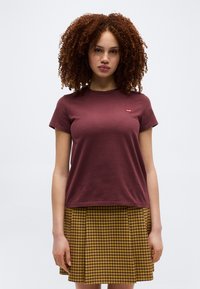 Νεαρή γυναίκα με σγουρά μαλλιά, φορώντας μπορντό κοντομάνικο μπλουζάκι και κίτρινη πλισέ φούστα με καρό σχέδιο, στέκεται μπροστά σε απλό φόντο.