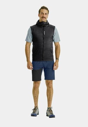 Black padded vest with high collar and zipper, worn over a light blue shirt, paired with navy shorts featuring black accents, and grey shoes.