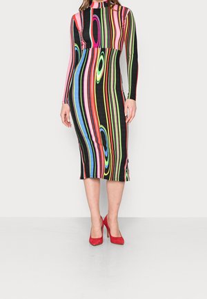 Kobieta w długiej, wielokolorowej sukience midi w paski z długim rękawem, w czerwonych butach na wysokim obcasie, stojąca na tle jednolitego tła.