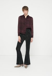 Donna che indossa una blusa a quadri scuri rossi trasparenti, pantaloni a campana neri con motivo e scarpe con tacco kitten beige, in piedi contro uno sfondo bianco semplice.