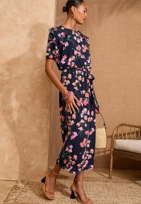 Cvjetna midi haljina u tamnoplavoj boji s ružičastim i narančastim cvjetovima, s volanima na rukavima, remenom oko struka i opuštenim krojem. Nositi s sandalama na remen.