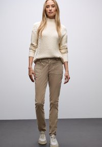 Pantalones de algodón beige con un corte ajustado, que presentan bolsillos laterales, combinados con un suéter de punto color crema con un patrón texturizado. Zapatillas blancas completan el conjunto.