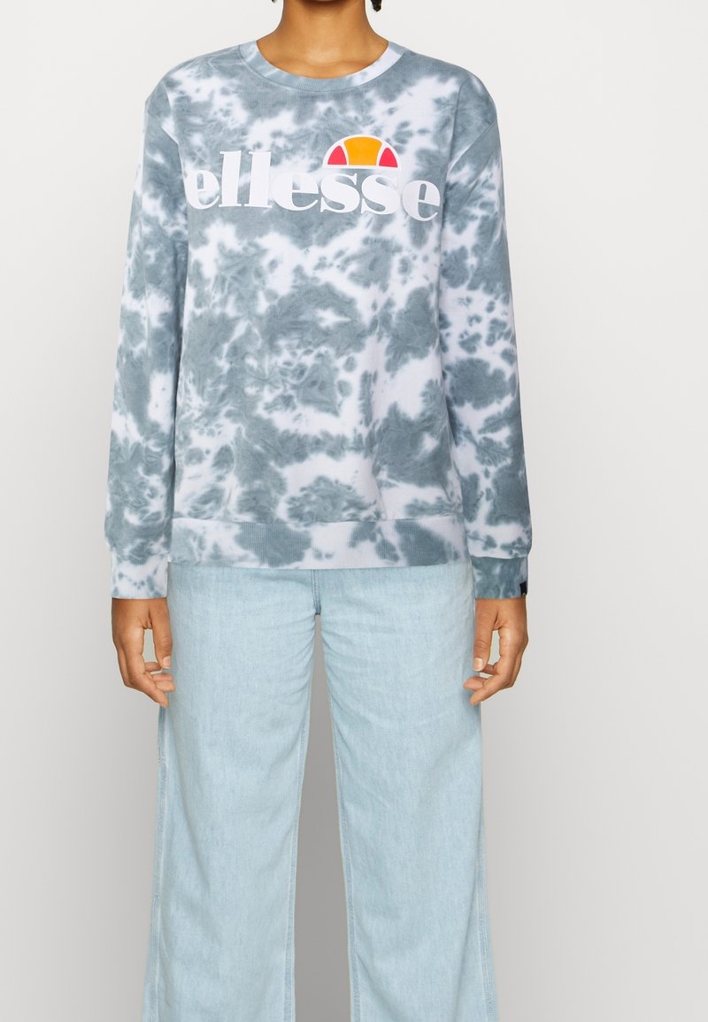 Personne portant un sweat-shirt tie-dye gris et blanc avec le logo « ellesse » en blanc et orange, associé à un jean large bleu clair.