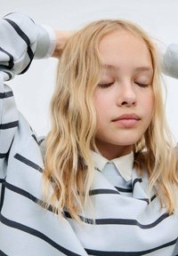 Pulover albastru deschis cu dungi orizontale negre, având o textură moale, purtat peste o cămașă albă cu guler. Părul este ondulat și blond.