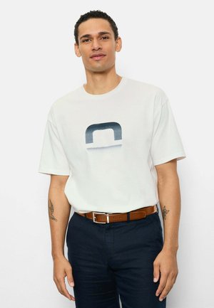 Man met kort krullend haar, gekleed in een wit T-shirt met een blauw logo, donkere broek, bruine riem en tatoeages op beide onderarmen, staand tegen een witte muur.