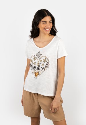 Junge Frau lächelnd, trägt ein weißes T-Shirt mit floralem Herzmotiv und dem Schriftzug "VIVA VIDA", kombiniert mit hellbraunen Shorts, steht vor weißem Hintergrund.