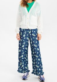 Biały dzianinowy kardigan z kieszeniami na zieloną koszulkę, zestawiony z granatowymi spodniami w kwiatowy wzór z falbaną na dole oraz jasnymi butami sportowymi.