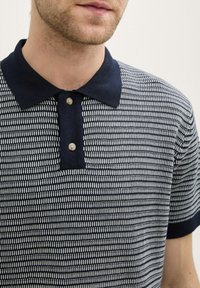T-shirt polo marine avec un motif texturé blanc, dotée d'une patte de boutonnage à trois boutons et de manches courtes. Les boutons sont ronds et de couleur neutre.