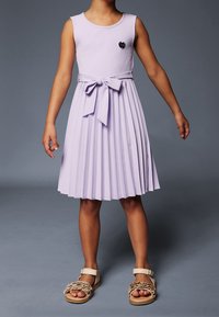 Dziecko w bezrękawowej, lawendowej sukience plisowanej z kokardą w talii i beżowych sandałach z falbanami, stojące na tle gładkiego, szarego tła.