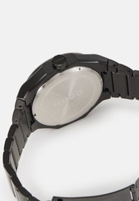 Black metal wristwatch with a textured bracelet, octagonal case, and a stainless steel back featuring engraved logo. Crown positioned on the side.