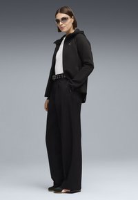 Donna che indossa una felpa nera con zip, maglietta bianca, pantaloni larghi neri con cintura a occhielli, scarpe slip-on nere e occhiali da sole su sfondo grigio.