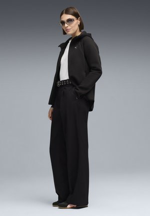 Kobieta w czarnej bluzie z zamkiem, białej koszuli, szerokich czarnych spodniach z paskiem z oczkami, czarnych wsuwanych butach i okularach przeciwsłonecznych na szarym tle.