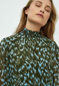 Grüne und blaue gemusterte Bluse mit hohem gerafftem Hals, strukturiertem Design und langen Ärmeln, die ein abstraktes Muster in kontrastierenden Farben aufweist.