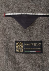 Grau strukturierter Stoff mit einem schwarzen Etikett, auf dem "Manteco" in Gold sowie ein Emblem und die Farben der italienischen Flagge dargestellt sind. Akzentstoff in Marineblau.