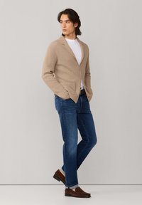 Muž v béžovom pletenom saku, bielom tričku, modrých džínsoch a hnedých mokasínach, stojaci s rukami v kapsách a pozerajúci sa do strany.