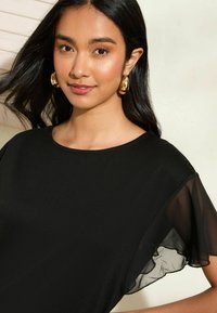 Černé šaty s krátkým rukávem, které mají volný průsvitný materiál na rukávech. Modelka vystavuje přirozený make-up na světlém pozadí.