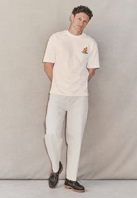 Maglietta bianca in cotone con stampa grafica, vestibilità comoda, abbinata a pantaloni larghi di colore chiaro e scarpe in pelle scura con dettagli in pizzo.