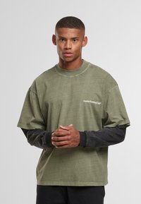 Noormees, kellel on lühikesed juuksed ja seljas oliiviroheline ning must kihiline pikkade varrukatega särgiga, hoiab käsi koos enda ees lihtsas taustal.