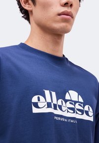 Νέος φοράει μπλε φούτερ ellesse με λευκό λογότυπο και τη φράση "PERUGIA ITALIA" στο στήθος.