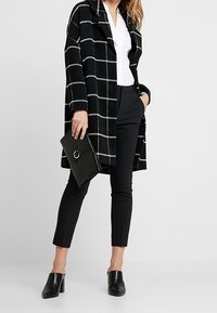 Schwarz-weißer karierten Mantel mit lockerer Passform, kombiniert mit schwarzen, engen Hosen und Absatzschuhen. Akzentuiert mit einer eleganten schwarzen Clutch.