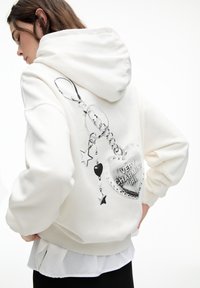 Sweat à capuche blanc avec un design graphique au dos, comprenant un pendentif en forme de cœur, des étoiles et le texte "FILLE TRÈS CHARMEUSE". Texture douce.