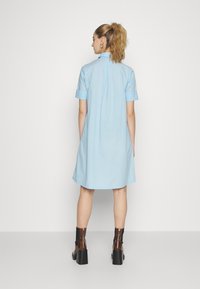 Vestido camisa azul claro, de mangas curtas, com uma bainha ligeiramente rodada e pregas nas costas, combinado com botas de cano baixo castanhas com salto.