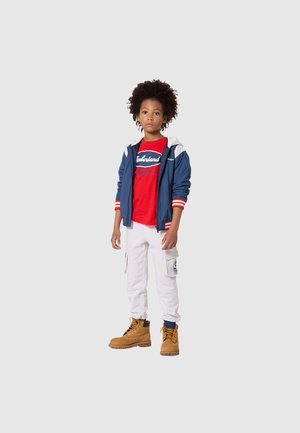 Veste bleue avec accents blancs et rouges, chemise rouge avec logo, pantalon cargo blanc et bottes couleur beige. Cheveux bouclés. Fond simple.