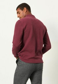 Bordeaux langärmliges Polohemd aus strukturiertem Stoff, mit einem Kragen und dezenten Rückennahtdetails, kombiniert mit grauen gemusterten Hosen.