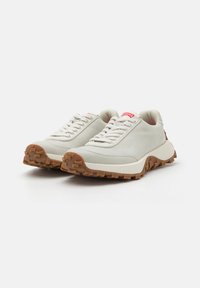 Light grey sneakers with textured surface, white laces, and chunky brown rubber sole. Features a logo on the tongue and sturdy design.