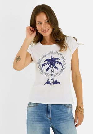 Femme aux longs cheveux bruns portant un t-shirt blanc avec un motif de palmier bleu et un jean bleu clair, souriante et se touchant les cheveux.